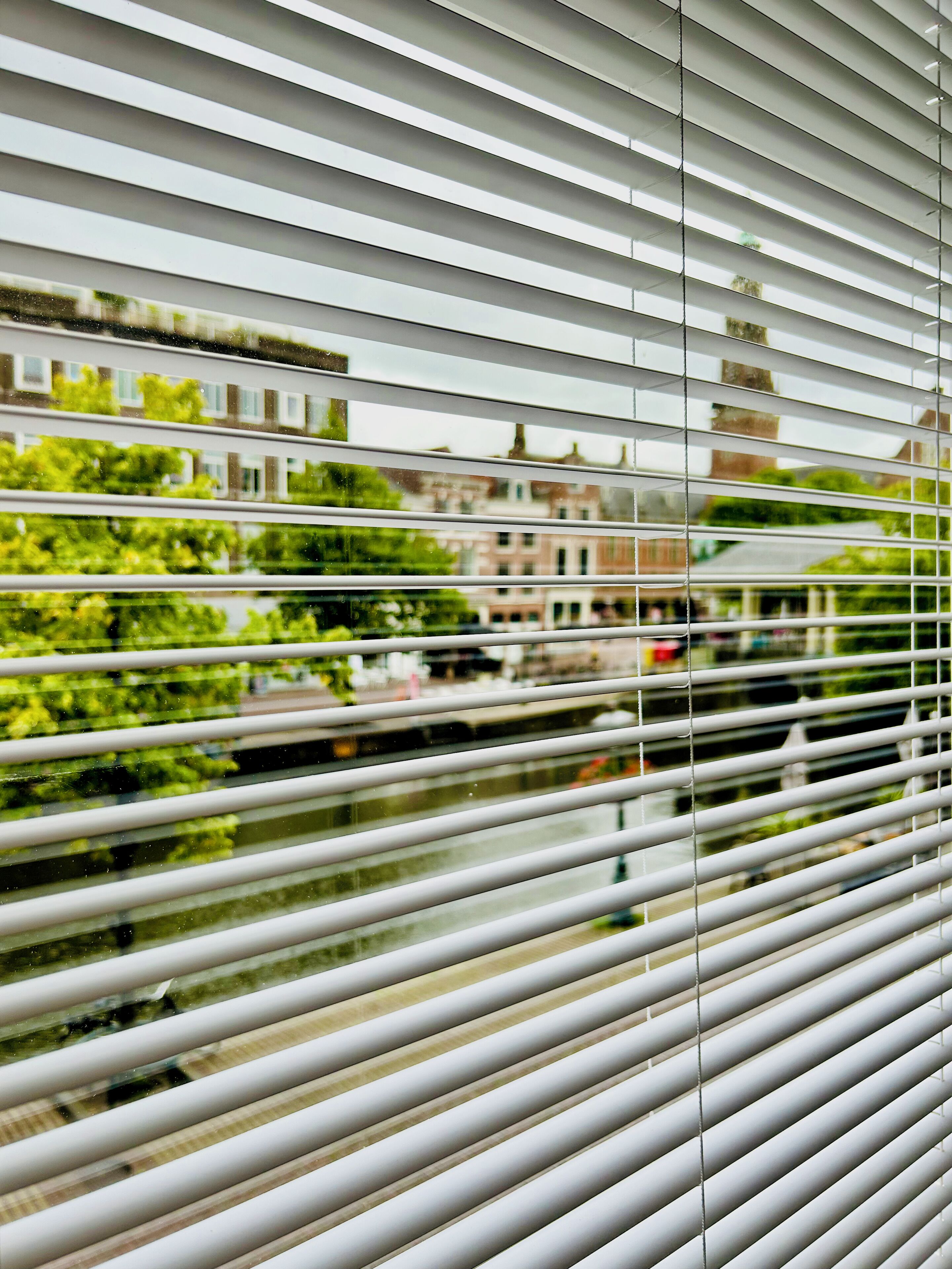 Uitzicht door jaloezieën met een reflectie van gebouwen en groen.