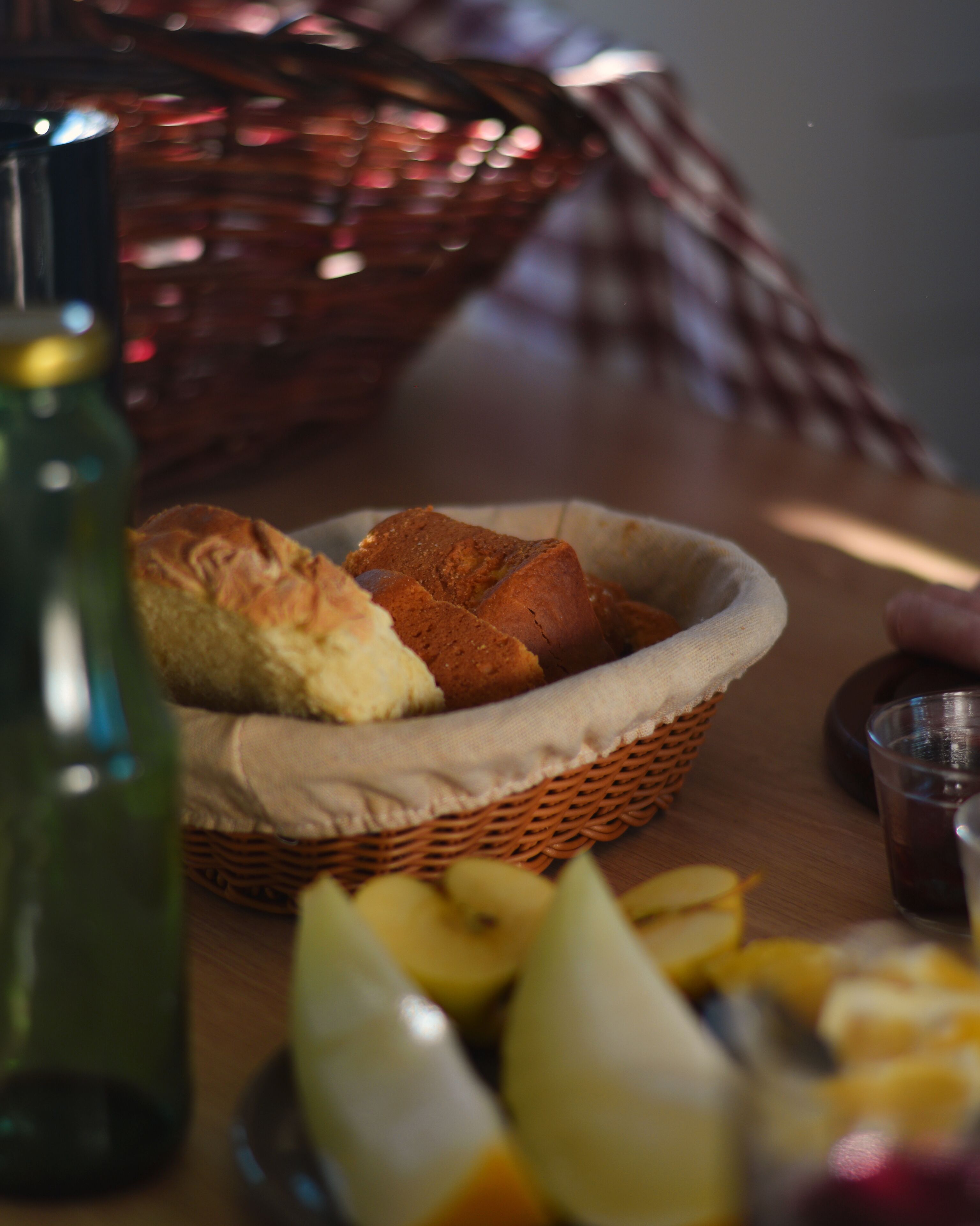 A wicker basket containing bread and a bottle on a wooden table.