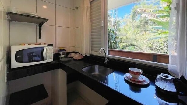 A compact kitchen with a microwave, sink, and window overlooking a lush garden. - Penedo (Itatiaia)