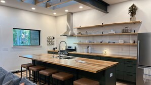 A modern kitchen with a central island, bar stools, a built-in oven, and a large window overlooking a forest.
