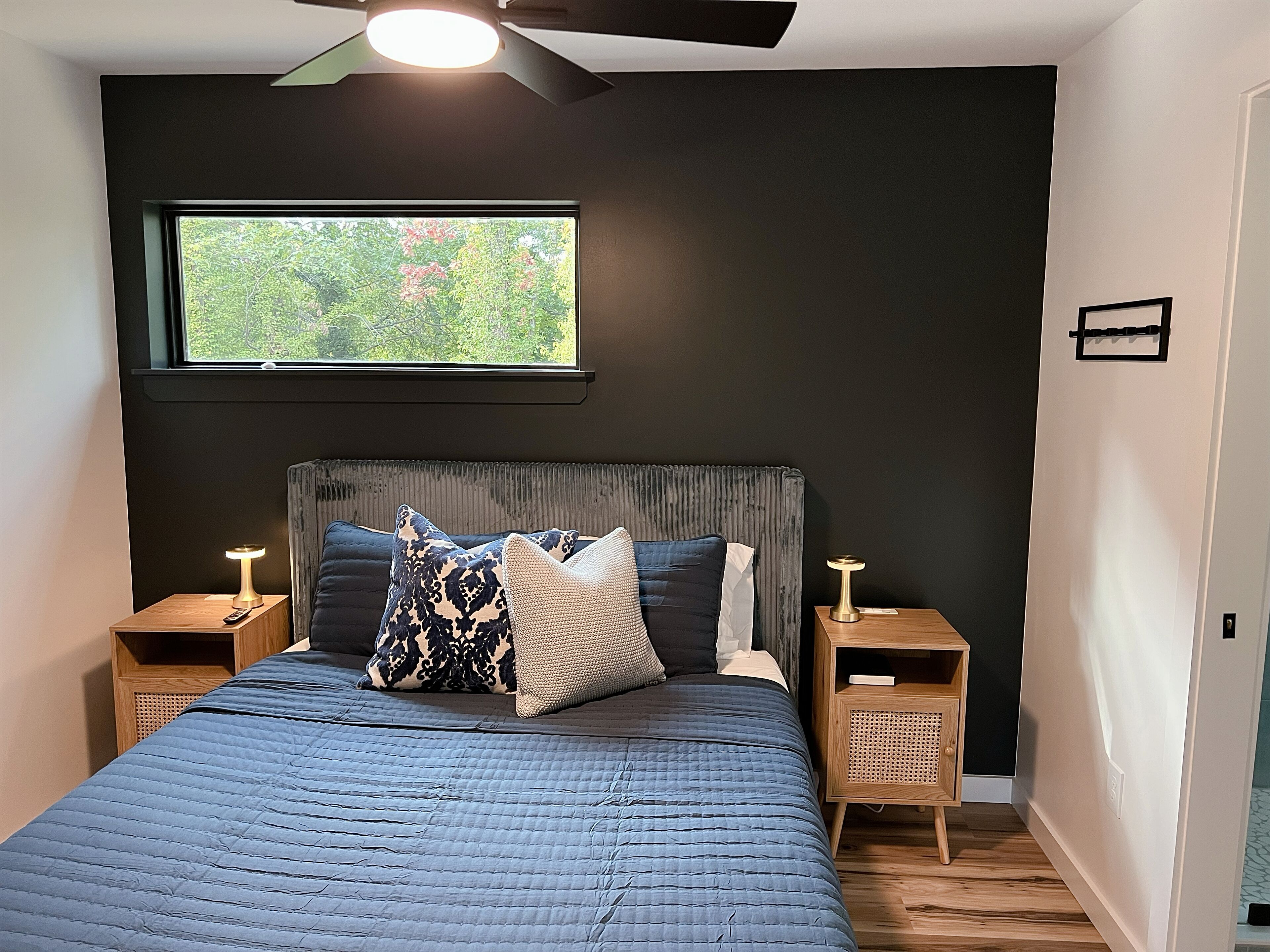 A bedroom with a dark wall, a large window, a bed with a blue quilt, and two bedside tables with lamps.