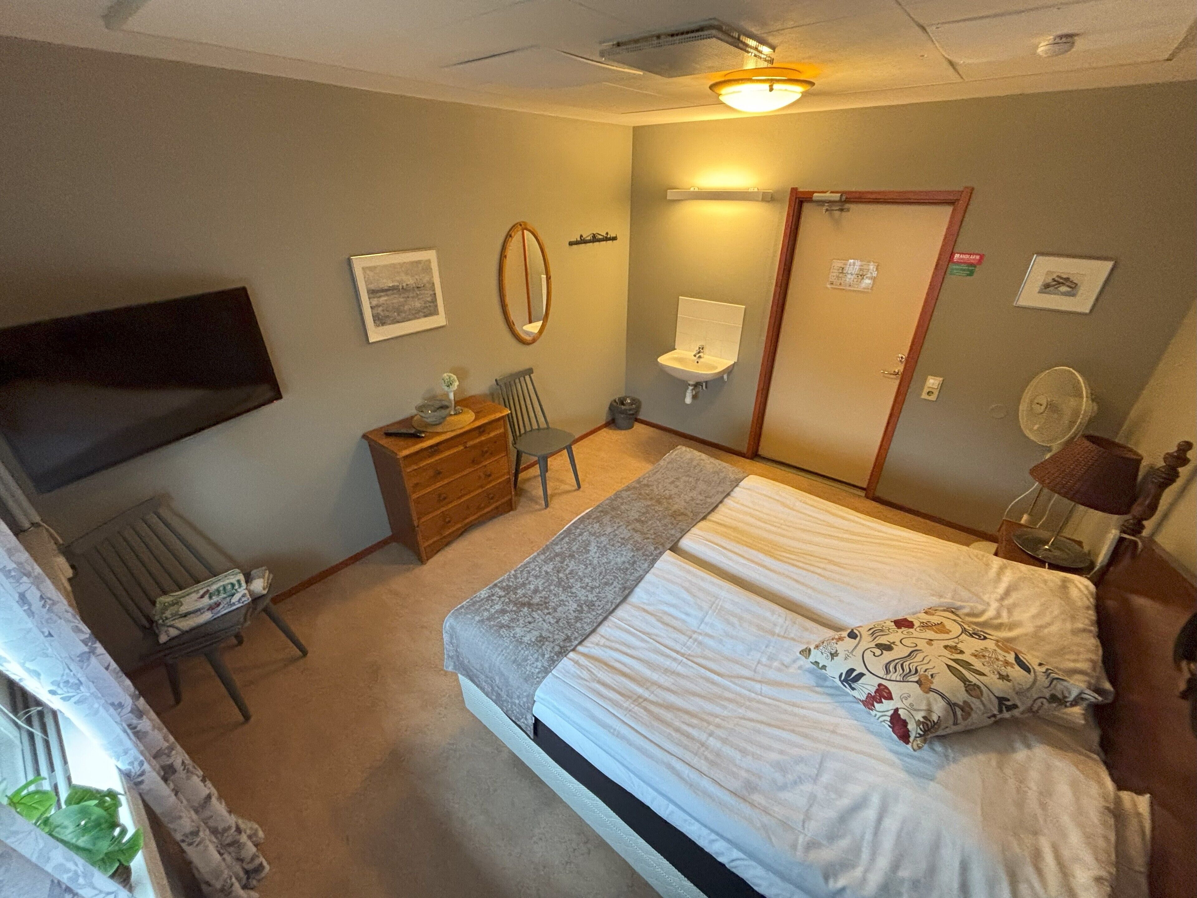 A bedroom with a bed, a dresser, a chair, a desk, a mirror, a radiator, and a window with curtains.