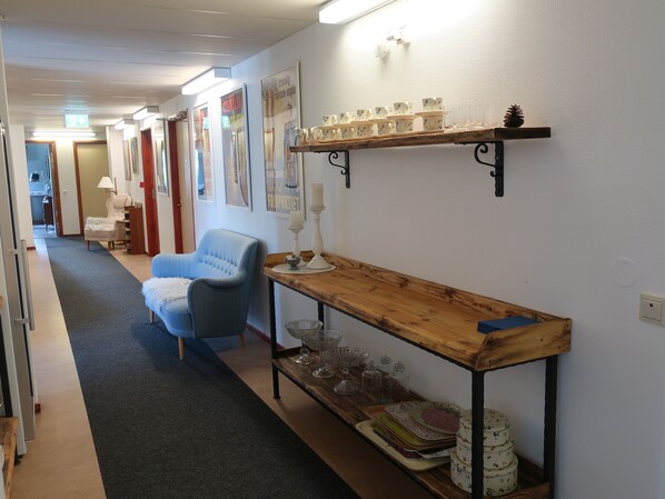 A hallway with a blue armchair, a wooden table with glassware, and wall-mounted shelves with decorative items.