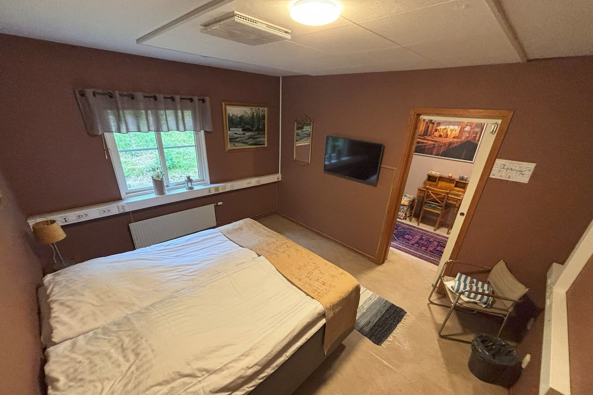 A bedroom with a bed, a window with curtains, a flat-screen TV, and a door leading to another room.