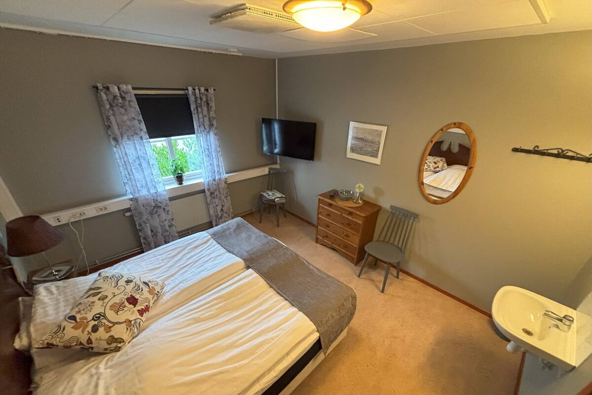 A bedroom with a bed, a dresser, a chair, a television, a mirror, and a window with curtains.