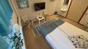 Un dormitorio con cama, televisor, silla, una mesita, una planta y baño con lavamanos y espejo.