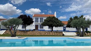 A two-story white building with blue trim and a tiled roof, surrounded by a swimming pool and trees.