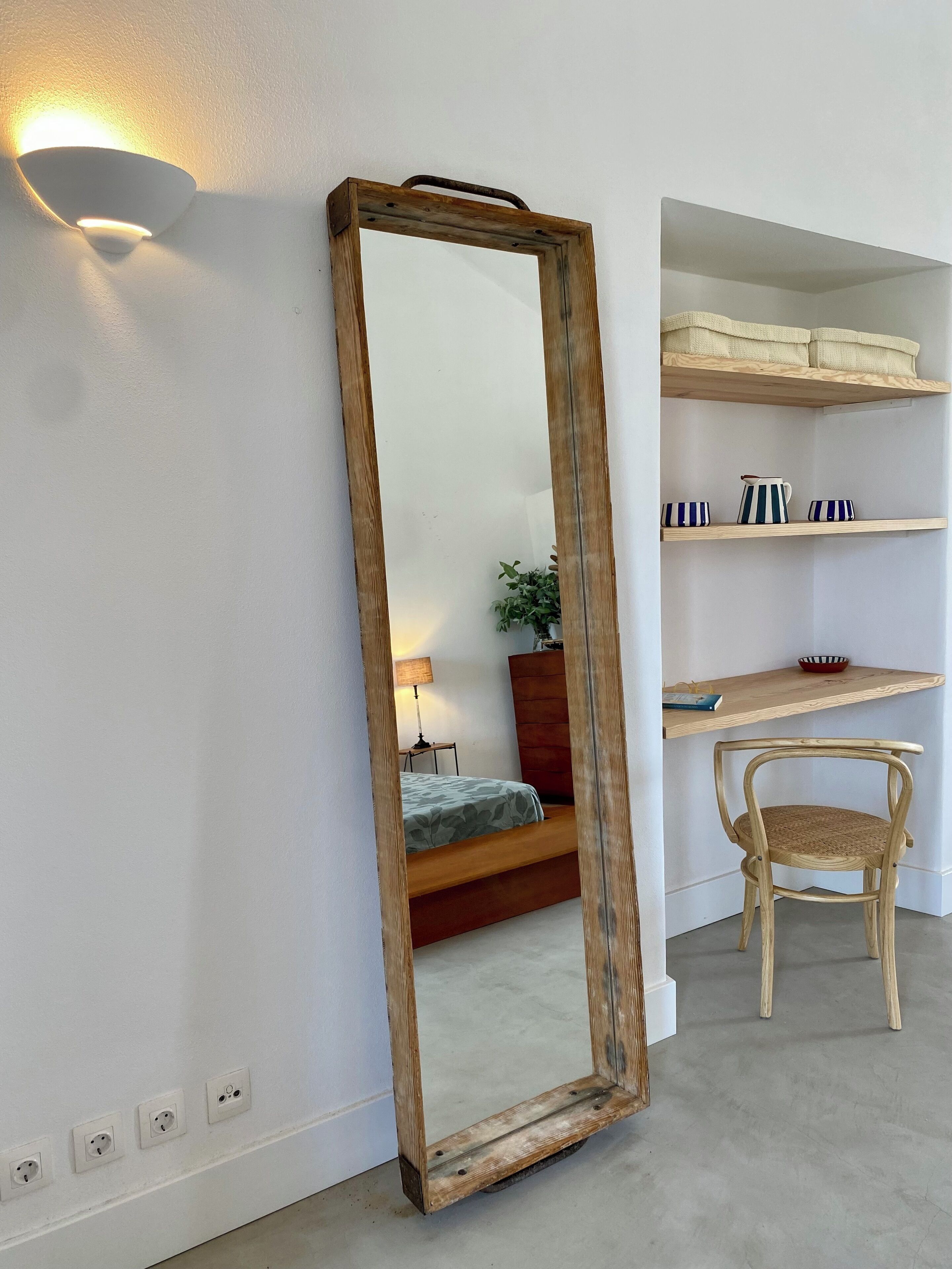 Una habitación con un espejo grande, una silla de madera, una cama, una mesita de noche, una lámpara y estantes con objetos decorativos.