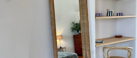 Una habitación con un espejo grande, una silla de madera, una cama, una mesita de noche, una lámpara y estantes con objetos decorativos.