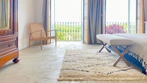 A bedroom with a large bed, a wooden dresser, a chair, and a balcony with a view of greenery.