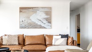 A living room with a brown leather sofa, a coffee table, and a wall-mounted painting of a beach scene. - Bicheno Blue Horizon - The family holiday house! (Glamorgan-Spring Bay)