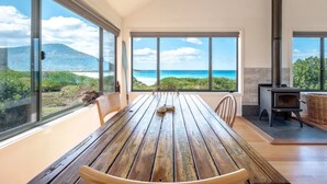 A spacious room with a large wooden table and chairs, large windows offering a view of the beach and ocean, and a wood-burning stove. - Silver Moon at Half Moon Bay - Paradise! (Glamorgan-Spring Bay)