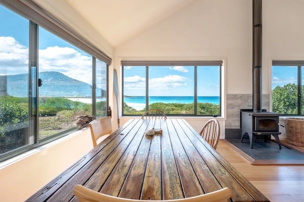 A spacious room with a large wooden table and chairs, large windows offering a view of the beach and ocean, and a wood-burning stove.
