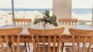 Une table à manger en bois, entourée de six chaises, un vase rempli de feuillage vert et une vue sur la plage et l’océan, visible à travers de grandes fenêtres.