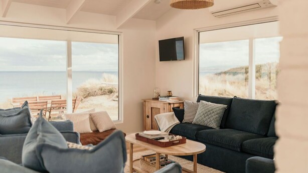 Un salon moderne doté d’une grande fenêtre donnant sur une plage, d’un canapé vert foncé, d’une table basse en bois et d’un téléviseur fixé au mur.