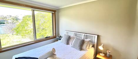 A neatly made bed with white linens and rolled towels, a bedside table with a lamp and decorative items, and a window with a view of greenery outside.