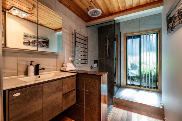 Ein modernes Badezimmer mit Holzboden, einem großen Spiegel, einem Waschbecken mit Seifenspender, einem Handtuchhalter und einer Duschkabine mit Glastür.