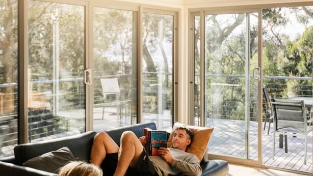 A person lying on a couch in a room with large glass doors leading to an outdoor deck.