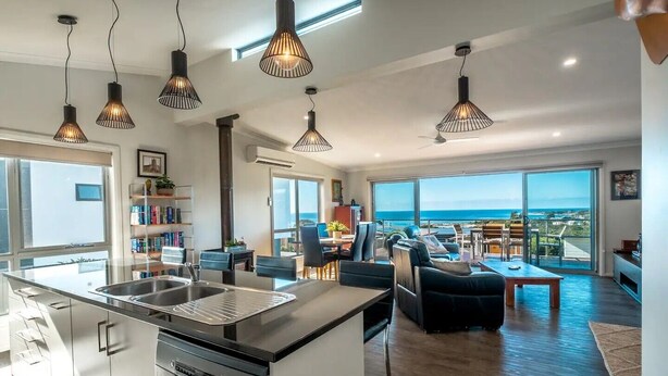 A modern kitchen with a central island, a dining area with a view, and multiple hanging pendant lights. - Ocean Sunrise - Beautiful views! (Glamorgan-Spring Bay)