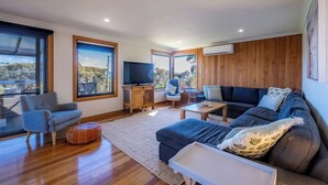 Amplia sala de estar con un sofá grande, un televisor sobre un mueble de madera y vistas al exterior a través de amplias ventanas.