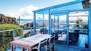 Un balcón con una mesa y sillas de madera, puertas de cristal y vista al mar y las colinas.