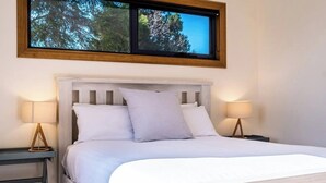 Una cama con ropa de cama blanca, dos mesitas de noche con lámparas, una ventana con vistas a árboles y un cabecero de madera.