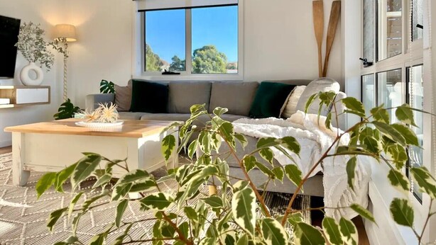 A modern living room with a large window, a sofa, a coffee table, and a television. - Viola - A coastal holiday retreat! (Glamorgan-Spring Bay)