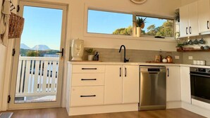 A modern kitchen with white cabinets, a stainless steel dishwasher, and a black faucet. There is a window with a view of mountains and a balcony with a white railing. - Viola - A coastal holiday retreat! (Glamorgan-Spring Bay)