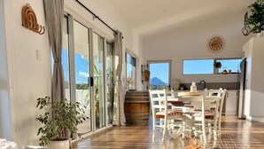 A spacious living area with a wooden dining table, a sofa, and a large window offering a view of the outdoors. - Viola - A coastal holiday retreat! (Glamorgan-Spring Bay)