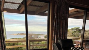 Un salón con un gran ventanal que ofrece vistas al mar y a las colinas. Hay un sofá de cuero negro con cojines estampados y una mesa de centro de madera.