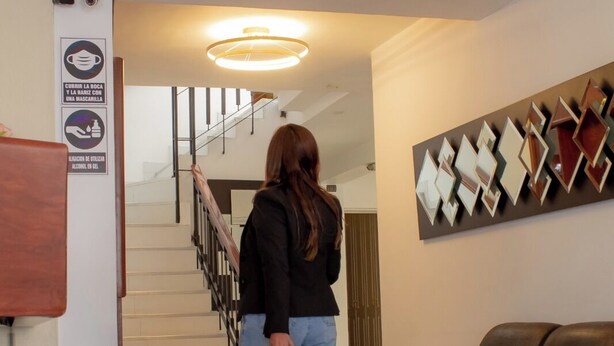 A woman with a suitcase in a modern lobby with a staircase, a wall-mounted mirror, and a brown leather couch. - Alsacia Boutique Hotel (Arequipa)