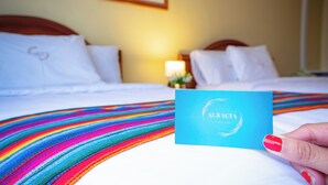 A hotel room with a bed, pillows, and a colorful striped blanket. A hand is holding a blue card with the text "ALSA CÍA". - Alsacia Boutique Hotel (Arequipa)