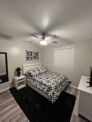 Une chambre à coucher avec un lit, une table de chevet, un ventilateur de plafond, une fenêtre et un téléviseur.