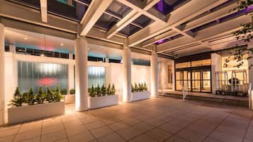 Amplio espacio interior con diseño arquitectónico moderno, que cuenta con pilares imponentes, piso de baldosas y fachada de vidrio que refleja luces.