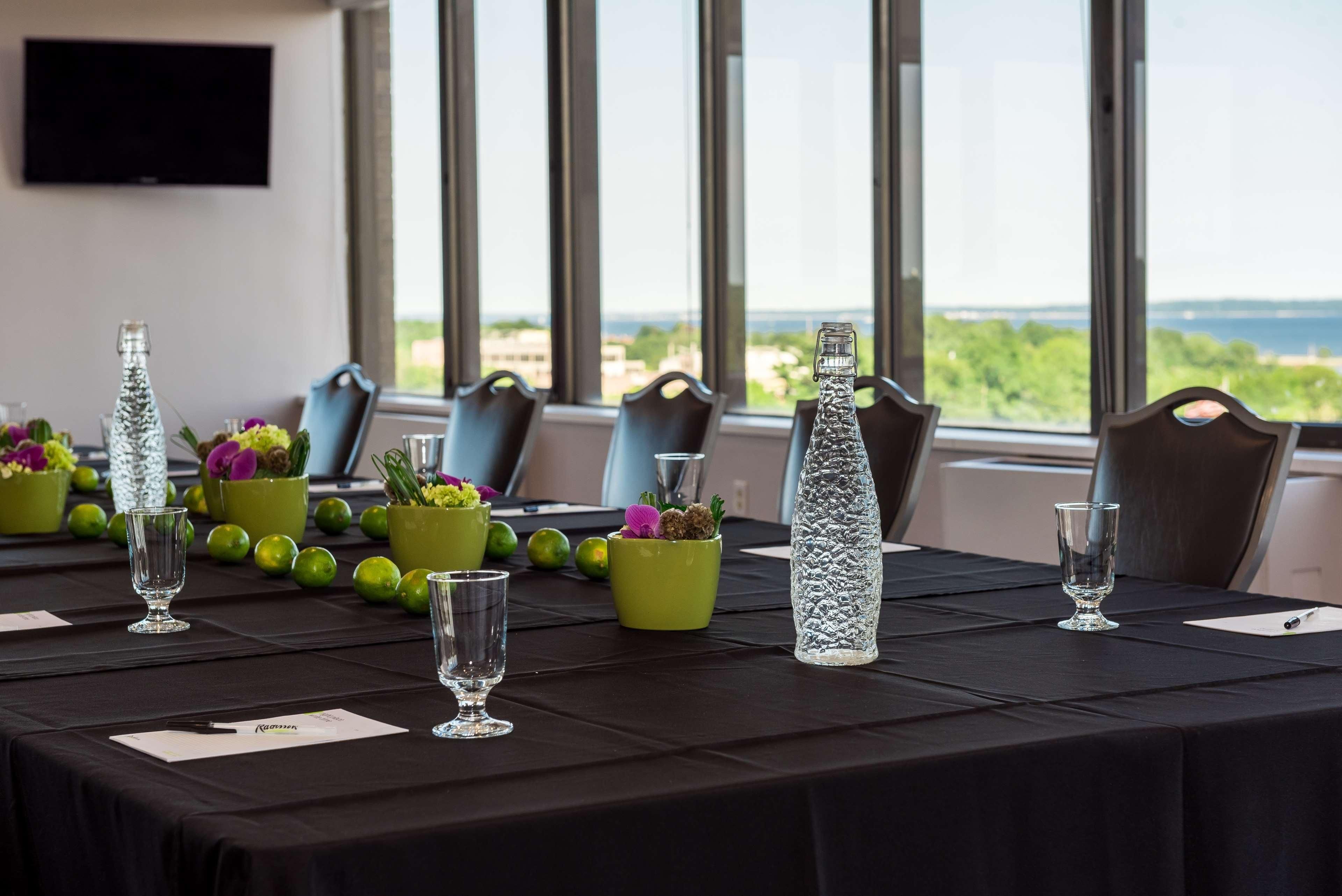 A conference table set with glassware, green vases with flowers, and a clear bottle, with a view of the sea through large windows.