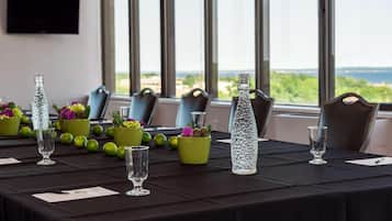 A conference table set with glassware, green vases with flowers, and a clear bottle, with a view of the sea through large windows.