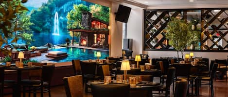 Un restaurante con un amplio ventanal que da a una cascada y piscina panorámicas, amueblado con mesas y sillas, y una pared decorativa con plantas.