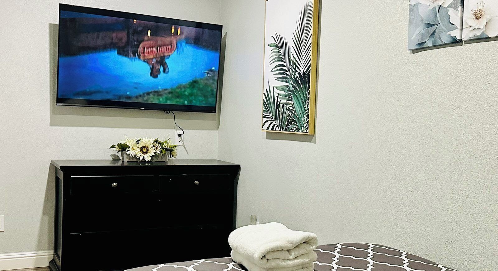 A room with a TV mounted on the wall, a black dresser with a vase of flowers, and a bed with a patterned pillow.