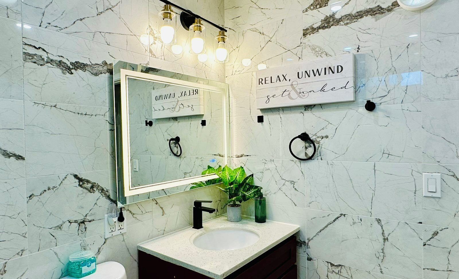 A bathroom with a marble backsplash, a sink with a black faucet, a mirror with a light fixture above it, and a sign that reads "RELAX, UNWIND get & naked".