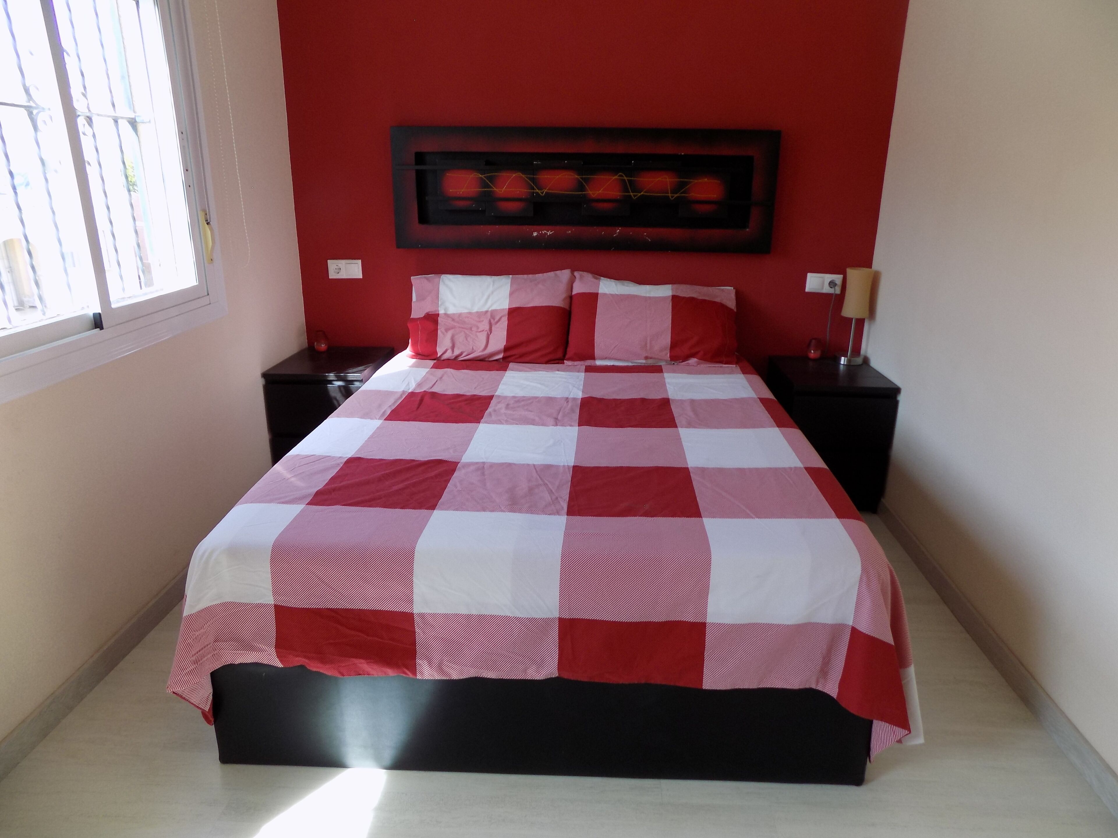 Un dormitorio con colcha a cuadros, una pared roja y una ventana con barrotes.