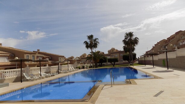 Une piscine entourée de bâtiments dotés de balcons et de palmiers, avec des chaises longues et des parasols à proximité.