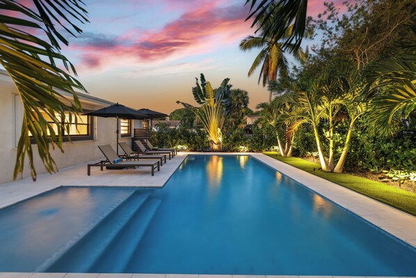 A swimming pool with lounge chairs and umbrellas, surrounded by tropical vegetation and a sunset sky.