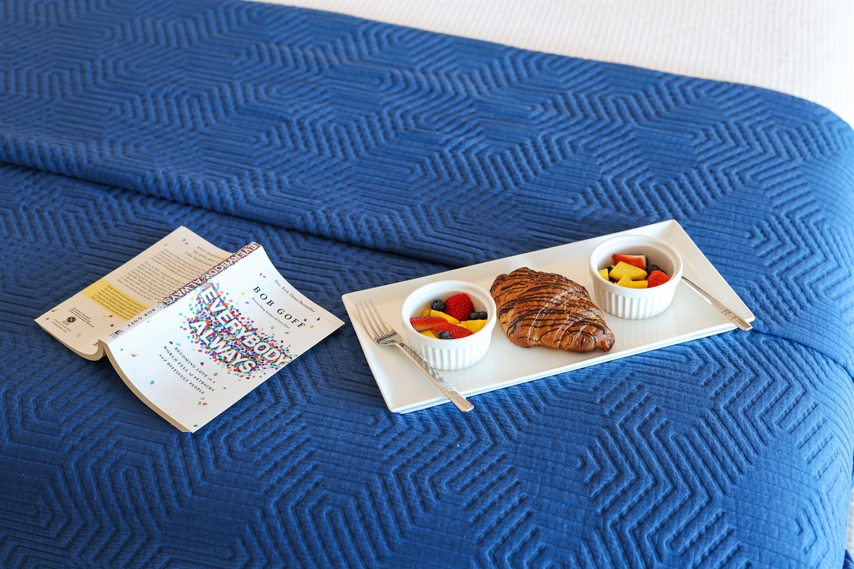 En seng med et blåt, struktureret betræk, en bog med titlen "The Book of Love" til venstre, og et bakke med en croissant og frugt til højre.