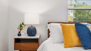 Ein ordentlich bezogenes Bett mit weißen, gelben und blauen Kissen, ein kleiner Nachttisch mit Lampe und eine Vase mit Blumen.