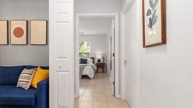 Ein Flur, der zu einem Schlafzimmer mit Bett, Nachttisch und Lampe führt. Im Vordergrund steht ein blaues Sofa mit einem gemusterten Kissen.