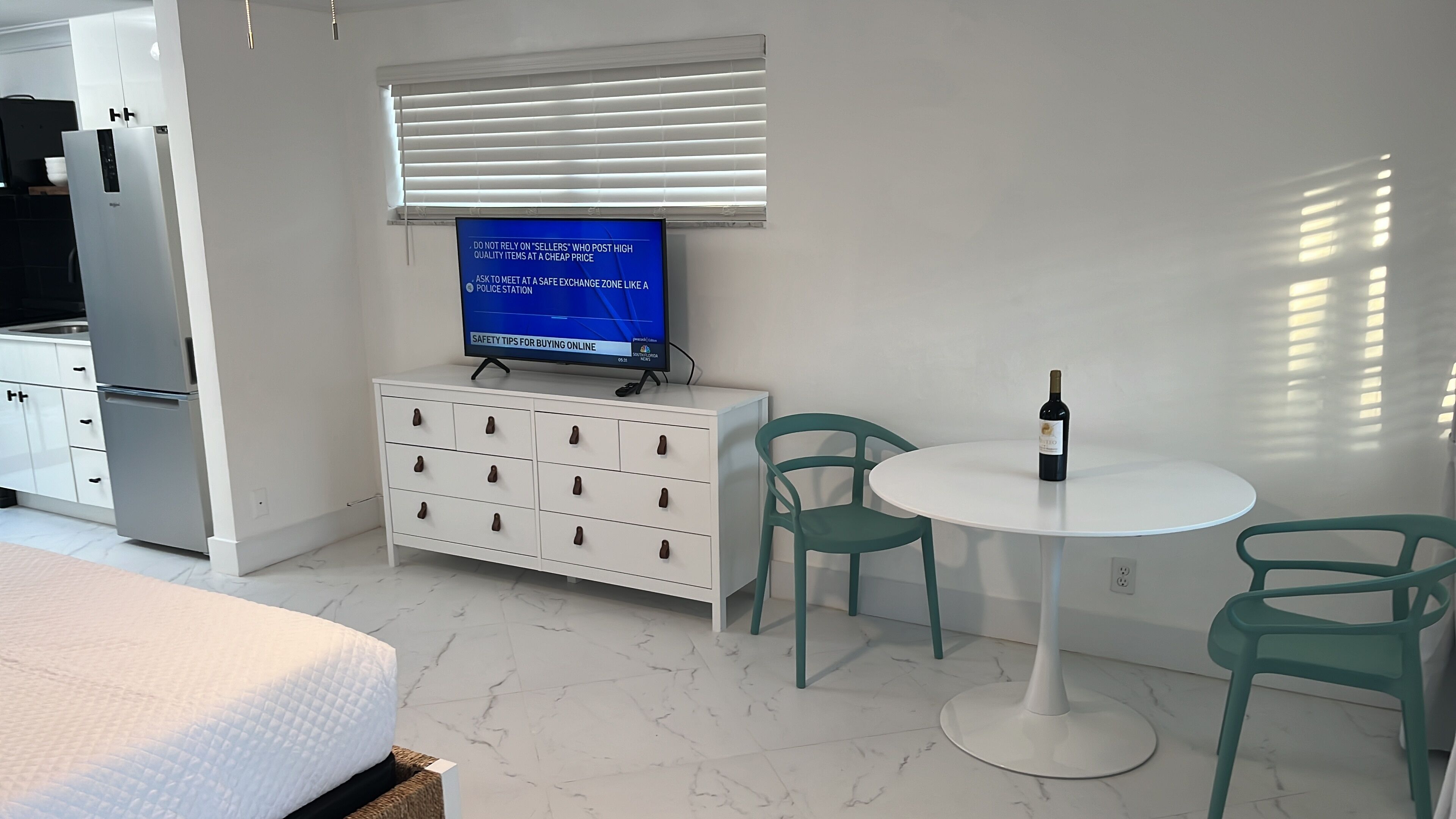 Een moderne hotelkamer met een bed, een witte dressoir, een televisie waarop veiligheidstips worden getoond, een klein rond tafeltje met een fles wijn en twee groene stoelen.