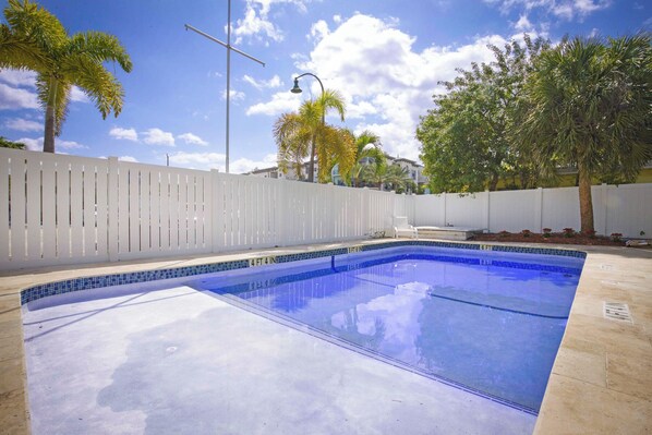 A swimming pool with a tiled edge, surrounded by a white fence, palm trees, and a street lamp. - La Mer Suites (Lauderdale-by-the-Sea)