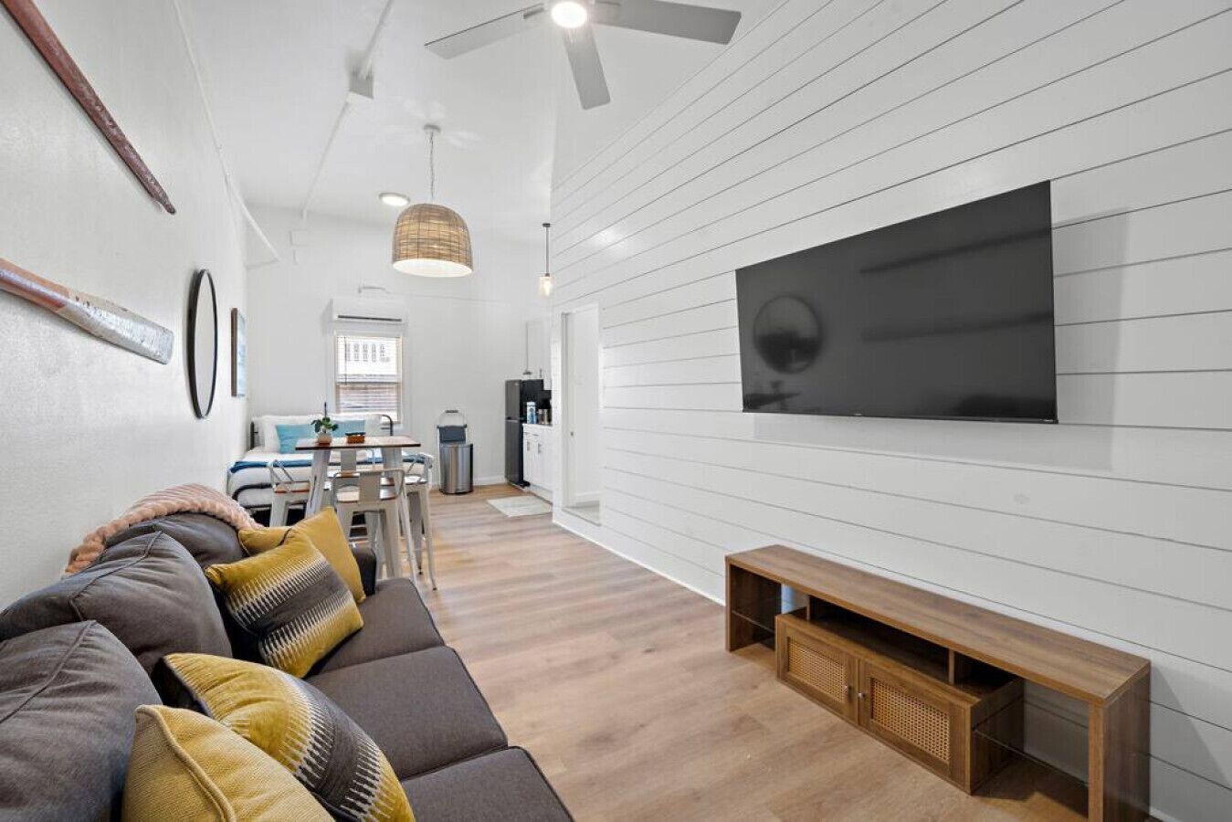 A modern living room with a large flat-screen TV, a sofa with cushions, a wooden coffee table, and a dining area in the background.