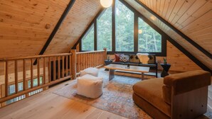 A cozy attic room with a wooden interior, a sofa, a coffee table, and a view of the forest through large windows. - Amazing Mountain Views + Hot Tub (Blue Ridge)
