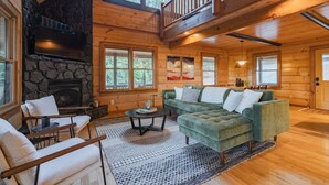 A cozy cabin interior with a stone fireplace, wooden walls, a green sofa, and a balcony. - Amazing Mountain Views + Hot Tub (Blue Ridge)